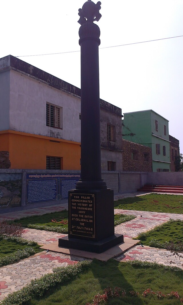 Picture showing an small plot with a lawn and paths for walking. A pillar stands in the middle. Behind the plot is another unknown building.